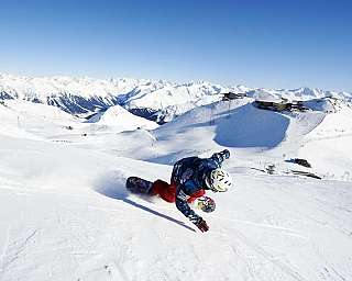 A snowboarder carving down a snowy mountain with peaks in the background.