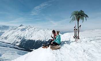 Two people sitting on snow with mountains in the background.