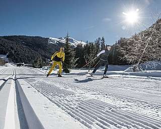 Zwei Skifahrer auf einer verschneiten Loipe bei Sonnenschein.