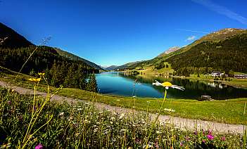 Bergsee mit klarem Wasser, umgeben von Wiesen, Wald und Bergen unter blauem Himmel