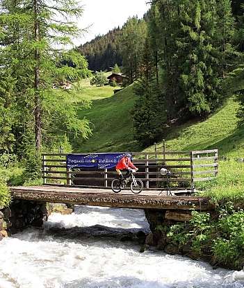 A cyclist crosses a bridge over a rushing river in a green landscape.