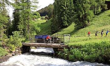 A cyclist crosses a bridge over a rushing river in a green landscape.