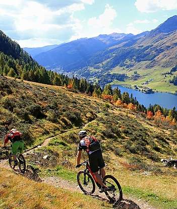 Two cyclists riding on a trail with mountains and a lake in the background.