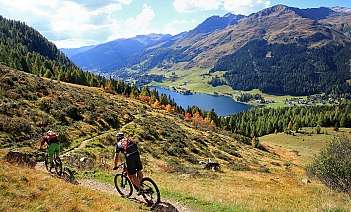 Two cyclists riding on a trail with mountains and a lake in the background.