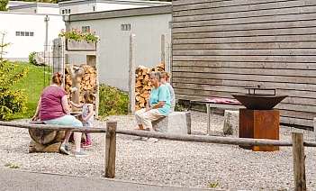 Drei Personen sitzen im Freien, umgeben von Natur und Holzstapeln.