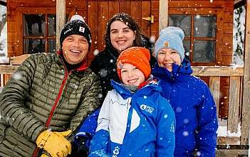 A family of four smiling in the snow near a wooden cabin.
