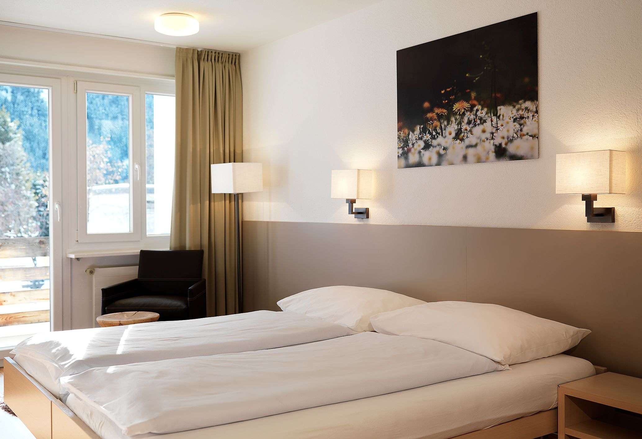 Helles Schlafzimmer mit Doppelbett, Fenstern, Vorhängen, Wandlampen und Blumenbild.