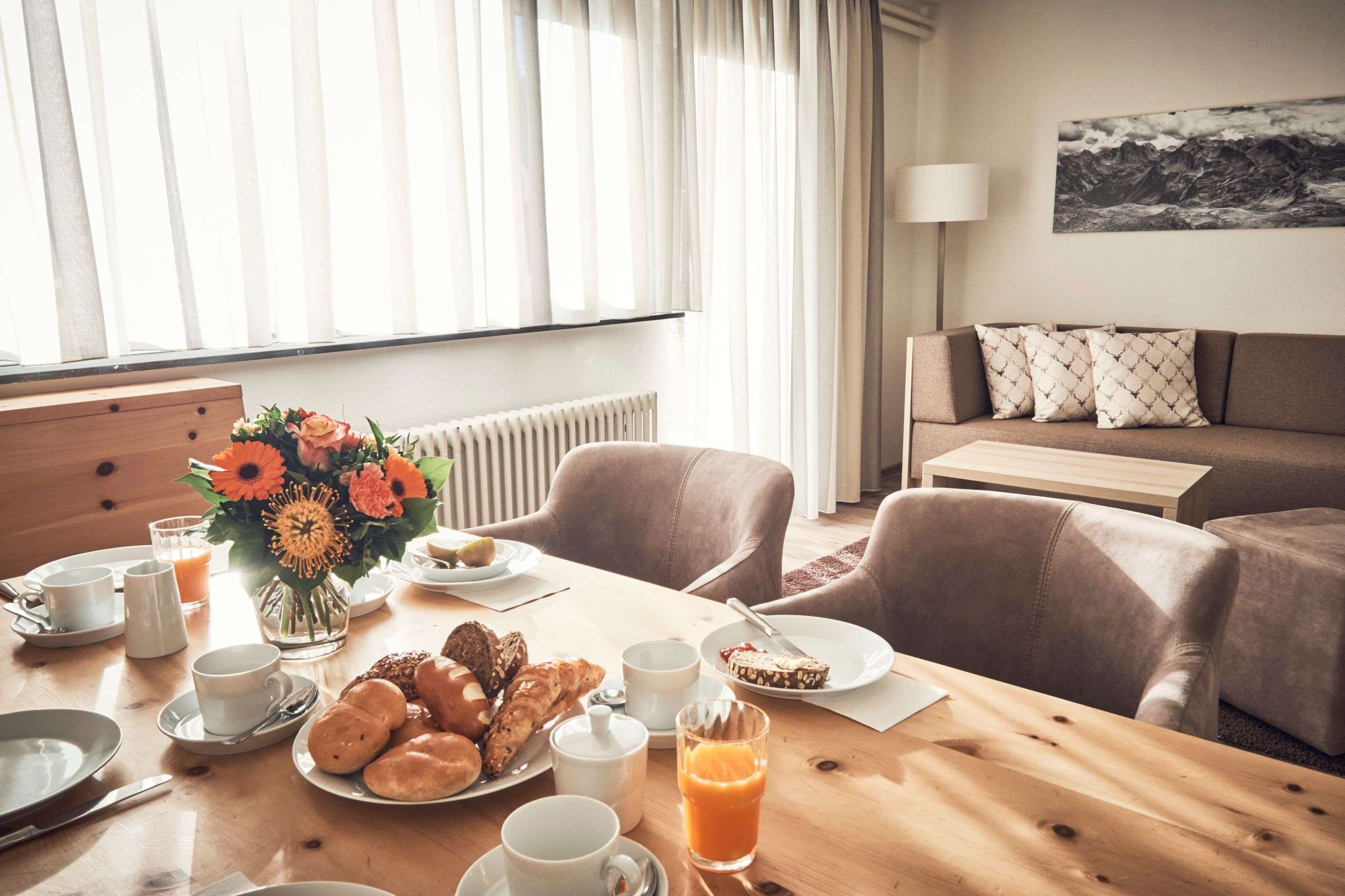 Ein heller Essbereich mit gedecktem Frühstückstisch, Blumen, Gebäck und Sofa im Hintergrund.